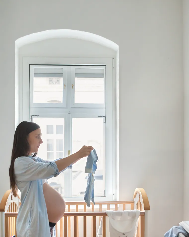 A pregnant woman folding baby clothes beside a crib